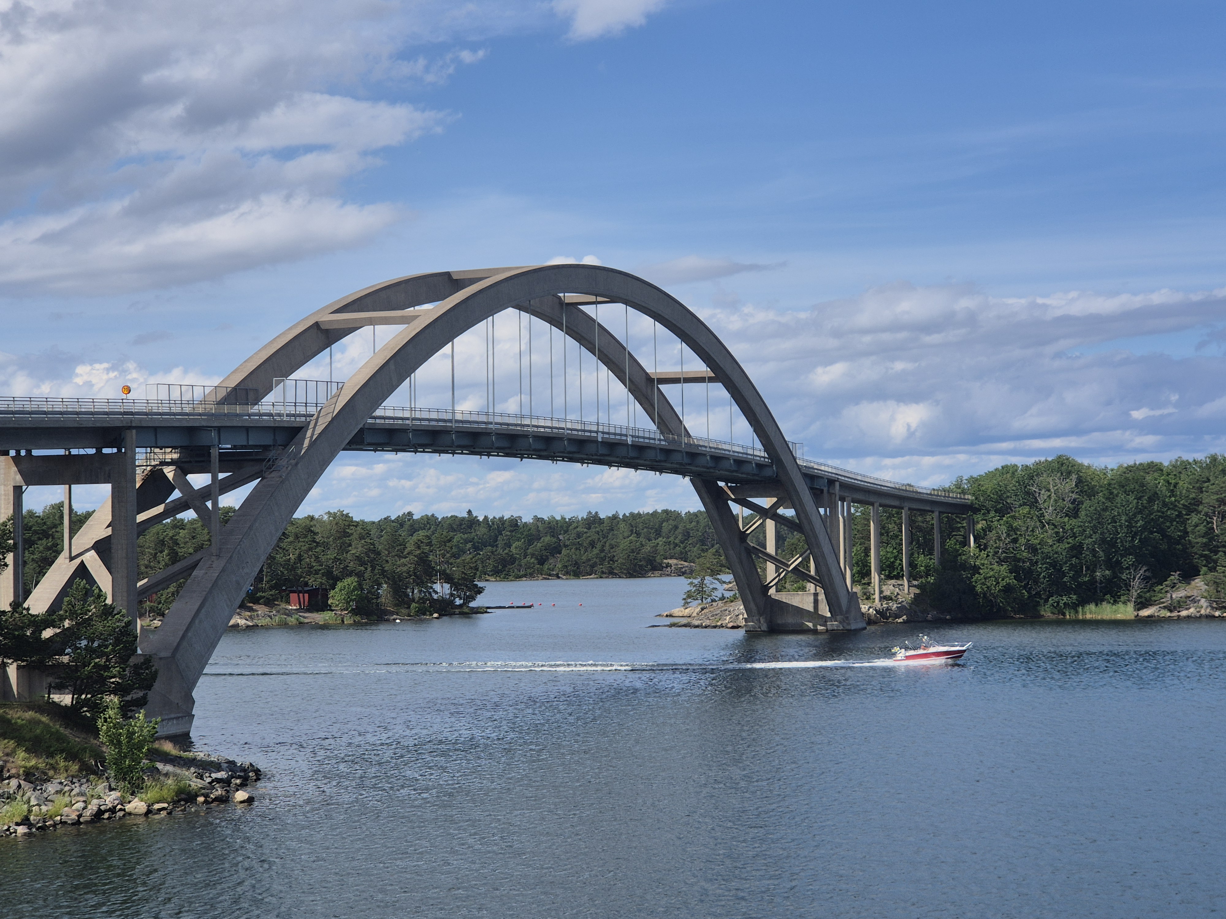 Djuröbron fotad från Stavsnässidan. röd motorbåt har precis kört under bron och svallvågor går hela